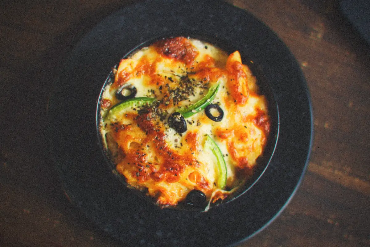 A bowl of baked pasta under bubbling browned cheese with black olives and green peppers.