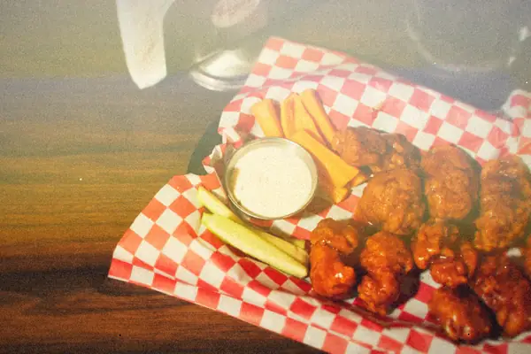 A basket of saucy buffalo wings on checkered paper with ranch dip, carrot sticks and celery.
