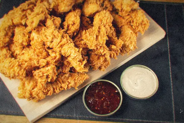 A board piled with crispy golden chicken tenders next to two small dipping sauces.
