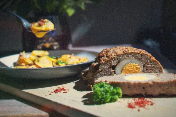 A sliced meatloaf on a wooden board with a plate of roasted potatoes behind it.