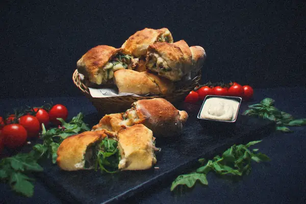 Small baked stuffed pizza pockets in a basket and on a slate board, with cherry tomatoes, arugula and a dip.