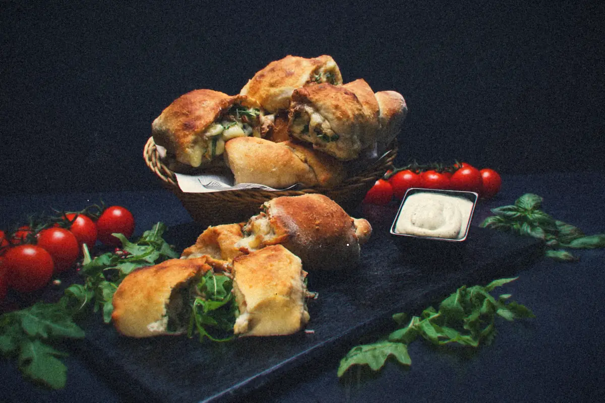 Small baked stuffed pizza pockets in a basket and on a slate board, with cherry tomatoes, arugula and a dip.