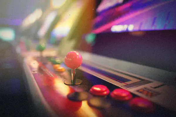 A pink ball-top joystick and red buttons on an arcade cabinet control panel, with more glowing cabinets blurred behind it.
