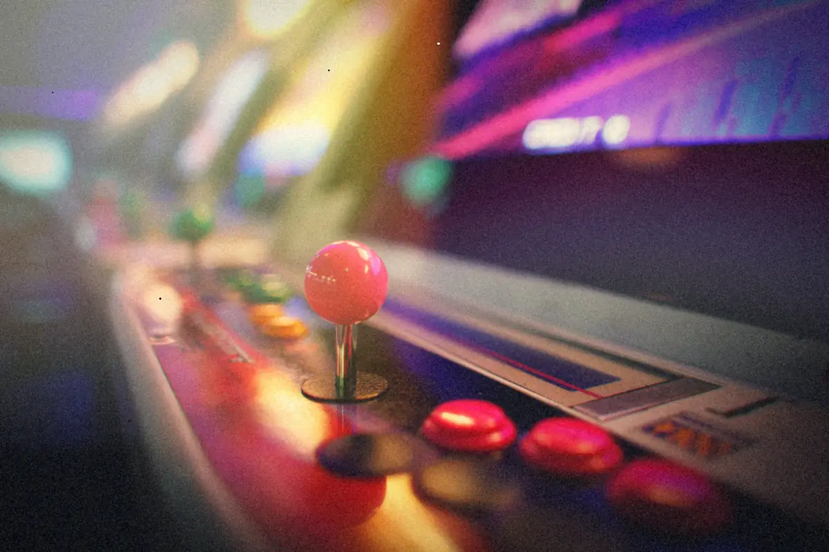 A pink ball-top joystick and red buttons on an arcade cabinet control panel, with more glowing cabinets blurred behind it.
