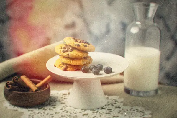 A stack of cookies on a small cake stand with fresh blueberries and a carafe of milk.