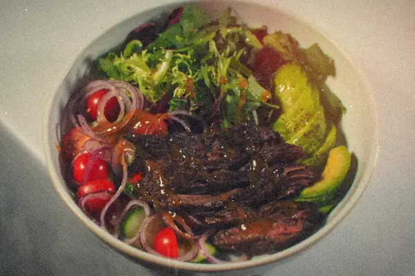 A salad bowl of sliced beef over greens with cherry tomatoes, red onion, cucumber and avocado.