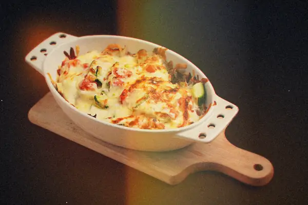 A cheesy baked vegetable casserole in an oval white dish on a wooden serving board.