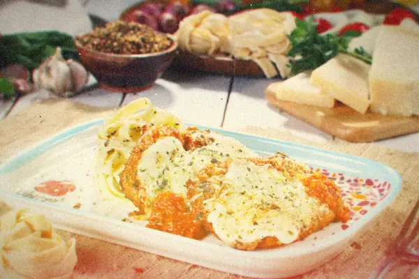 Breaded chicken parmesan with melted cheese and tomato sauce beside a nest of pasta, parmesan wedges behind it.