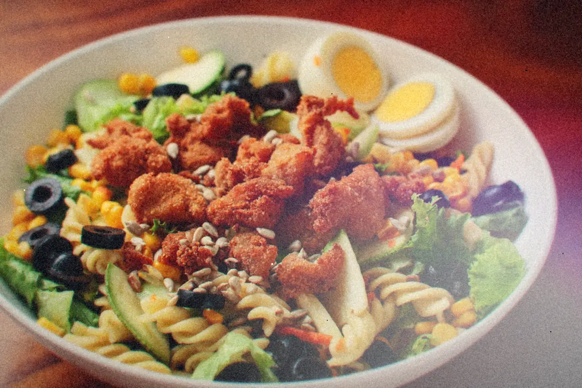 A bowl of rotini pasta salad with crispy chicken, olives, corn, lettuce and boiled eggs.