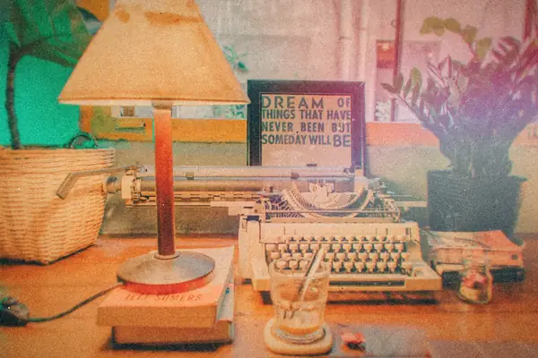 A vintage typewriter on a desk beside a table lamp and houseplants.