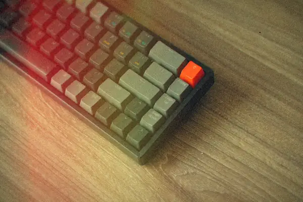 A gray mechanical keyboard with one orange key, on a wooden desk.