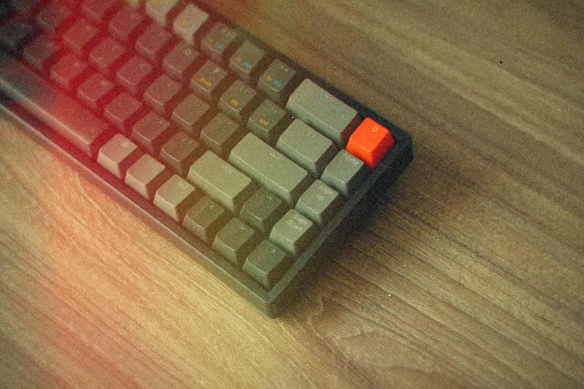 A gray mechanical keyboard with one orange key, on a wooden desk.