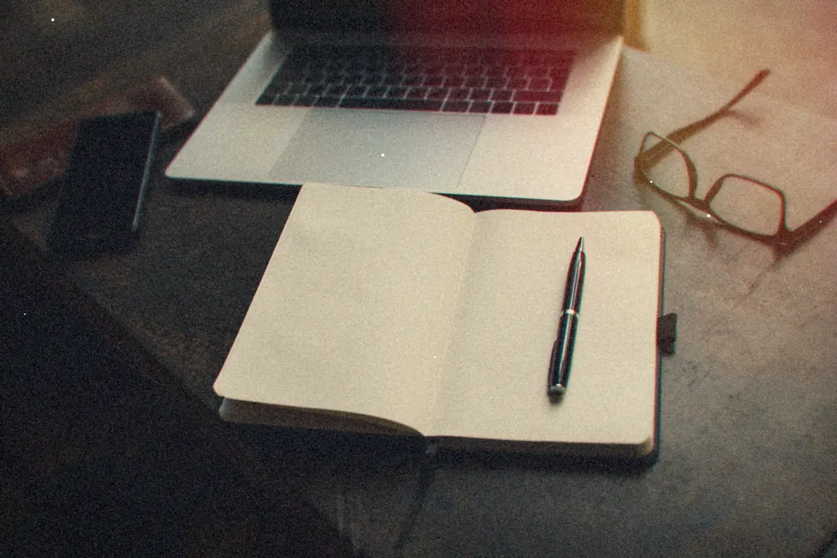 An open notebook with a pen beside a laptop, a phone and a pair of glasses on a dark desk.