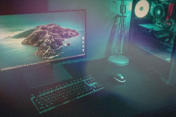 A dark gaming desk with a monitor, backlit keyboard and a glowing tower PC.