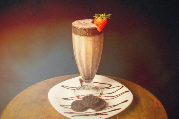 A tall chocolate cookies-and-cream shake with a strawberry on the rim, sandwich cookies on a chocolate-drizzled plate.