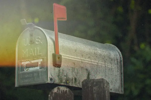 A classic metal U.S. mailbox with its red flag raised against green trees.