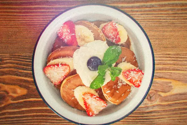 A bowl of small pancakes topped with cream, strawberries, banana slices, a blueberry and mint.