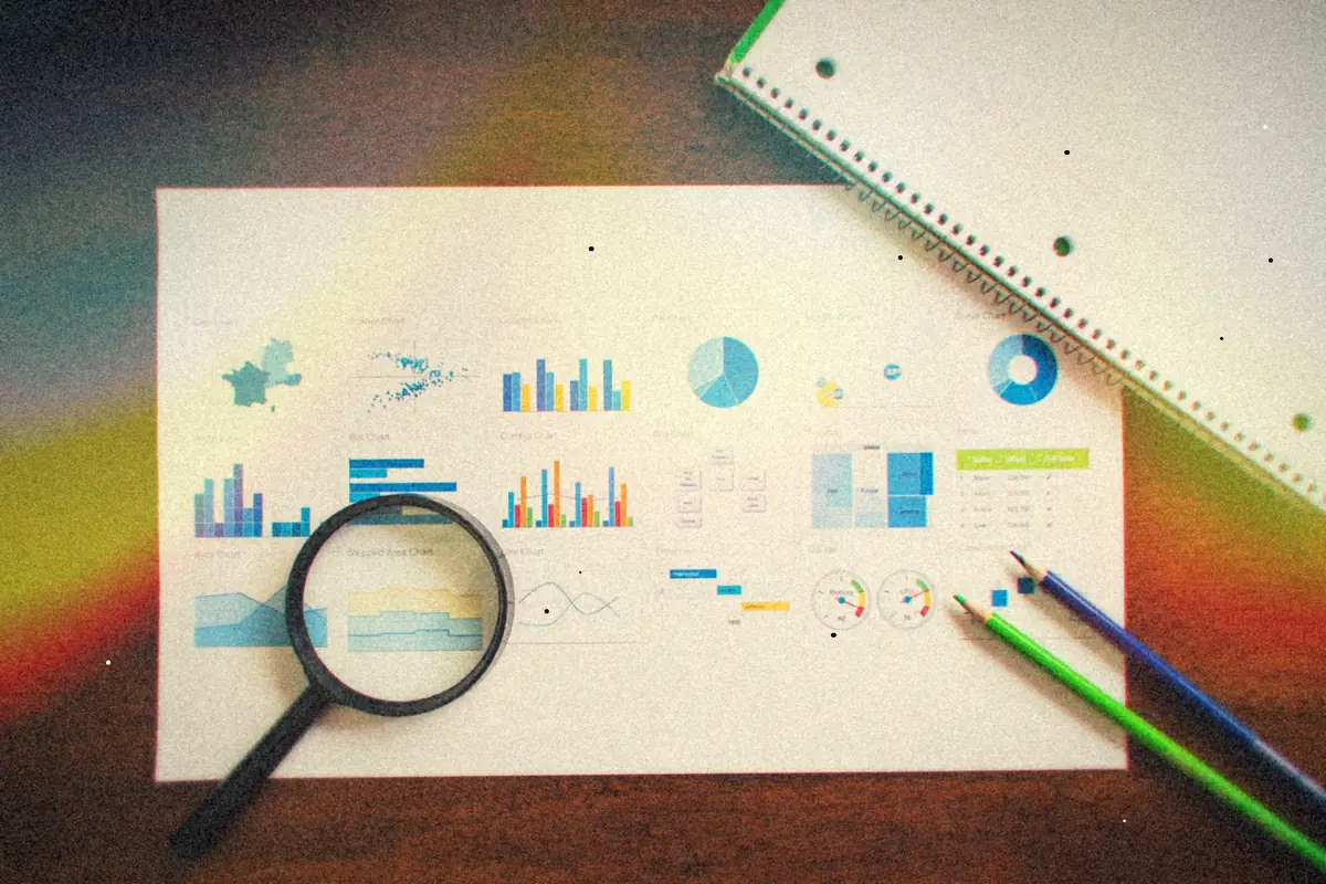 Printed bar and pie charts on a wooden table with a magnifying glass, pencils and a notebook.