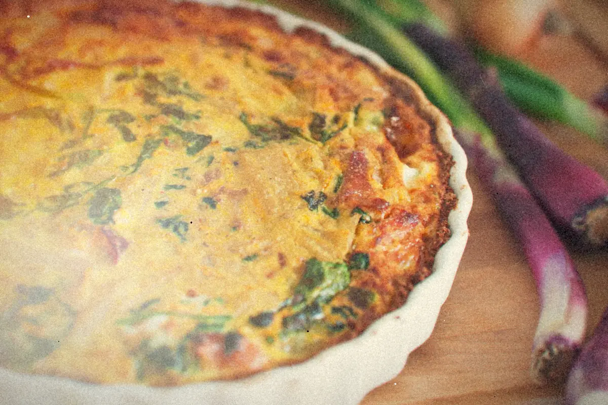 A baked egg casserole with spinach and onions in a white fluted dish beside red spring onions.