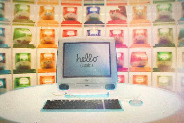 A single all-in-one Apple computer on a round white table in front of a wall of colorful iMacs.