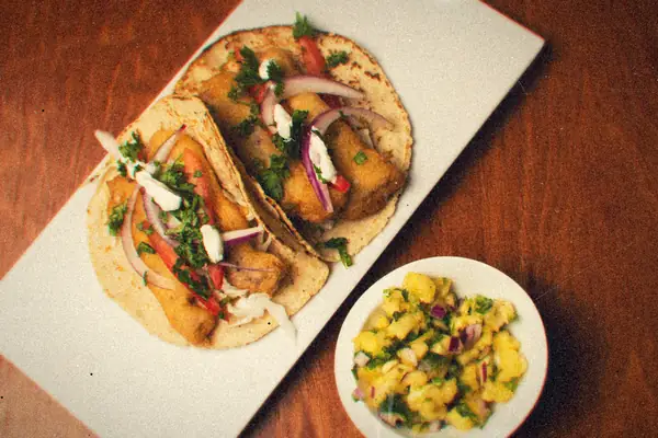 Two battered fish tacos with onions, cilantro and cream on a white platter, with a bowl of pineapple salsa.
