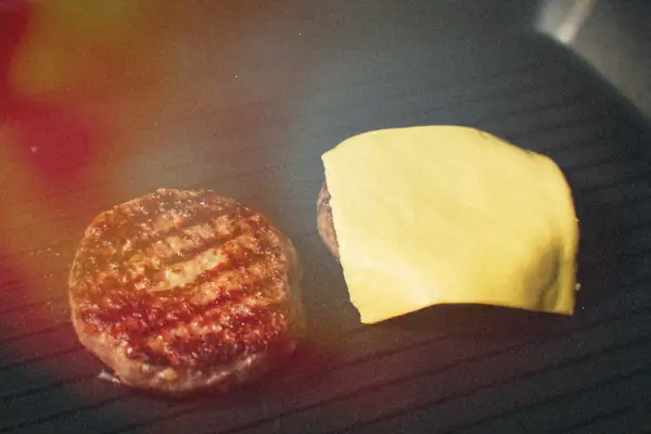 Two grilled beef burger patties on a grill pan, one topped with a slice of melting cheese.
