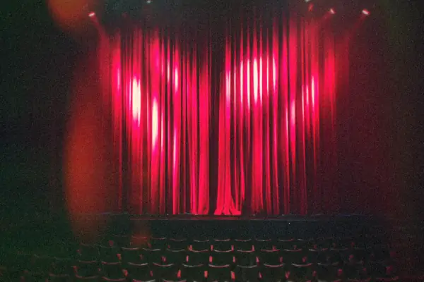 An empty theater with a red stage curtain lit by spotlights above rows of seats.