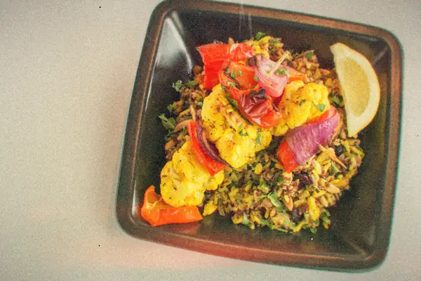 Roasted cauliflower, red peppers and red onion over herbed grains with a lemon wedge in a dark square dish.