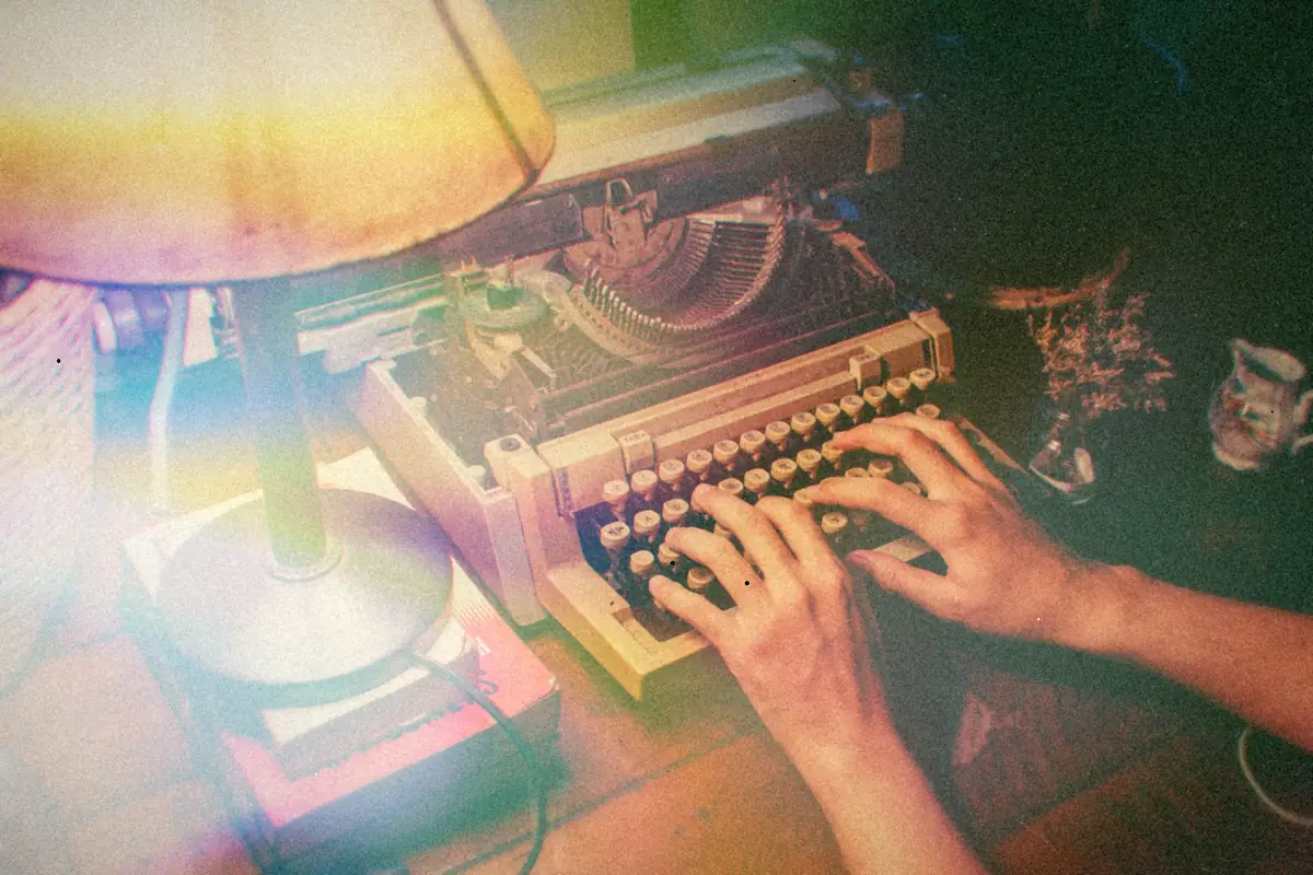 Hands typing on a cream typewriter under the warm glow of a table lamp.