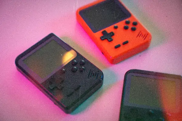 Three small off-brand retro handhelds in black and orange scattered on a pink-lit surface.