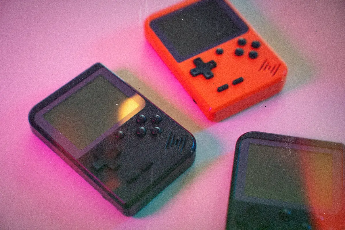 Three small off-brand retro handhelds in black and orange scattered on a pink-lit surface.