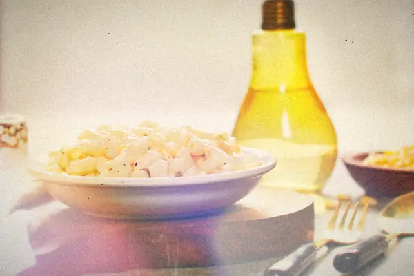 A white bowl of creamy elbow macaroni with corn on a wooden board, with forks and a bottle of oil nearby.