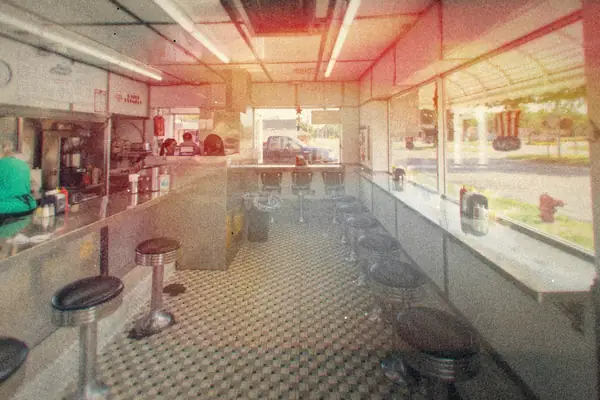 Inside an empty chrome diner with a long counter, stools and a checkered floor.