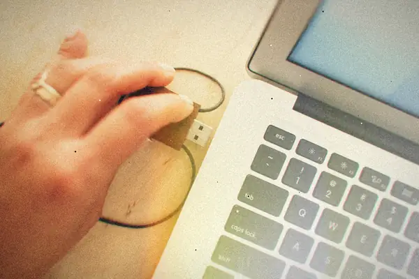 A hand plugging a wooden USB flash drive into the side of a silver MacBook.