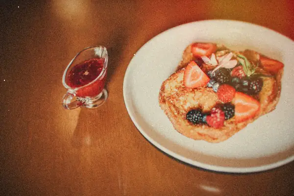French toast topped with fresh berries on a white plate next to a small pitcher of berry syrup.