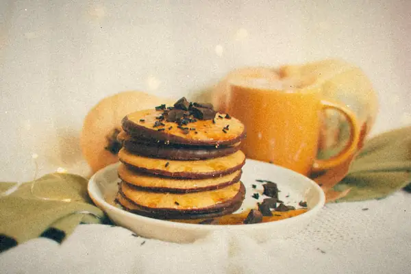 A stack of pumpkin pancakes with chocolate shavings beside a mug and whole pumpkins.