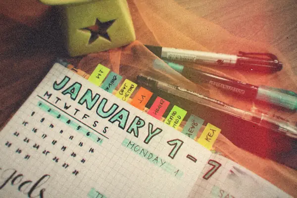 A paper weekly planner with goals and colorful tabs next to pens on a wooden desk.