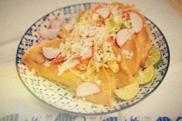 Folded crispy tacos topped with shredded lettuce, crumbled cheese and radish slices, with lime wedges on a blue patterned plate.
