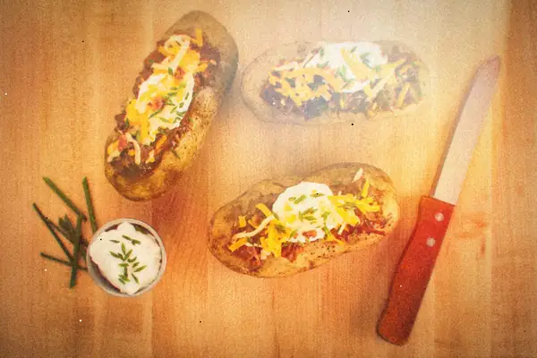 Three loaded baked potatoes with shredded cheddar, sour cream and chives on a wooden board.