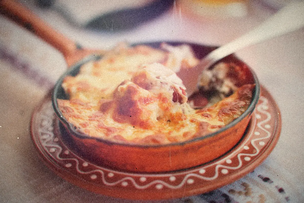 A cheesy baked meat dish with a browned, bubbly top in a clay pan, with a spoon lifting out a portion.