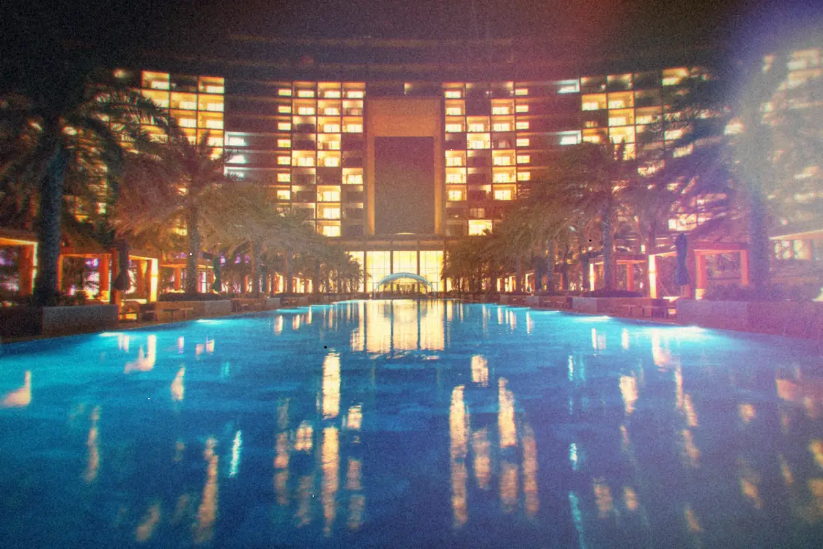 A long swimming pool lit up at night, lined with palm trees.