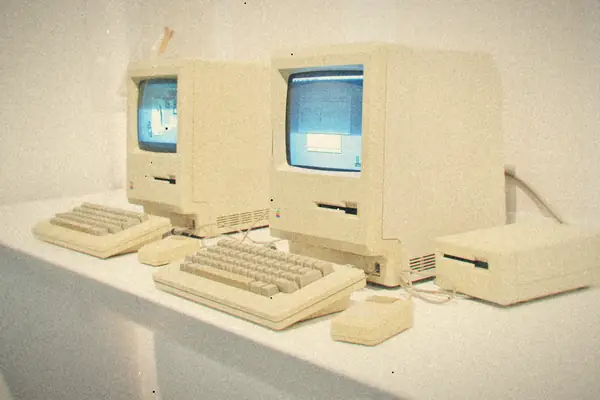 Two classic beige Macintosh computers with keyboards and a mouse on a white display table.