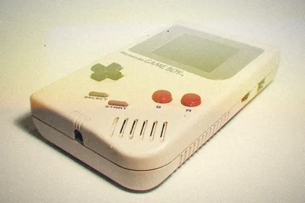 An original grey Game Boy lying on a white surface, shown at an angle with its d-pad and red A and B buttons.