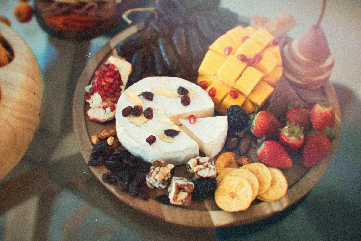 A round wooden board of brie, cheddar cubes, strawberries, dried fruit, nuts and banana chips.