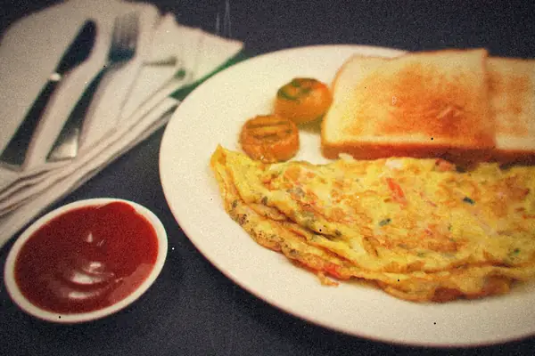 A folded omelet on a white plate with toast, grilled tomato and a dish of ketchup.