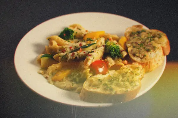 Creamy penne with broccoli, squash, peppers and a cherry tomato, served with garlic bread on a white plate.