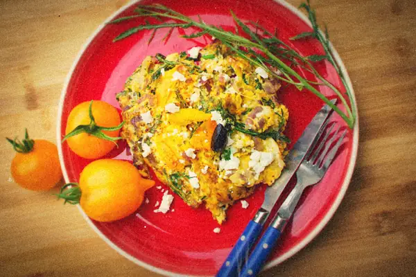 A vegetable frittata with crumbled feta and fresh herbs on a red plate, yellow tomatoes on the side.