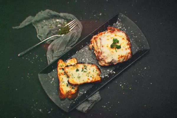 A cheesy baked tomato-sauce dish topped with parsley next to two slices of cheesy garlic bread on a black platter.