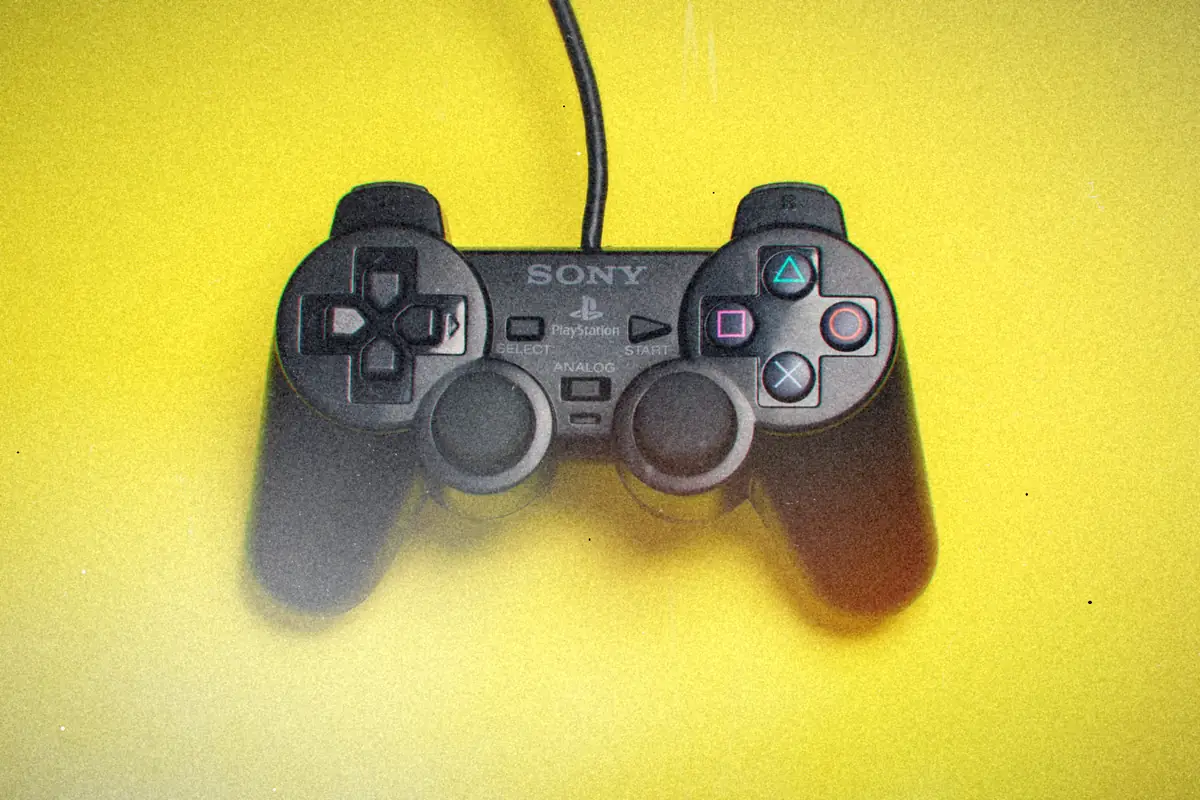 A black PlayStation 2 DualShock 2 controller, cable attached, lying on a bright yellow background.