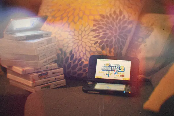 A stack of Nintendo DS game cases on a sofa with a silver DS on top and a 3DS XL beside them.